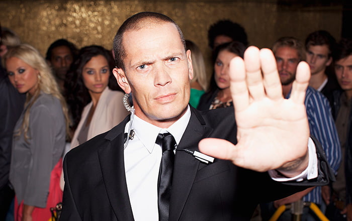 A security guard dressed in a suit holding up his hand to block entrance with partygoers in the background